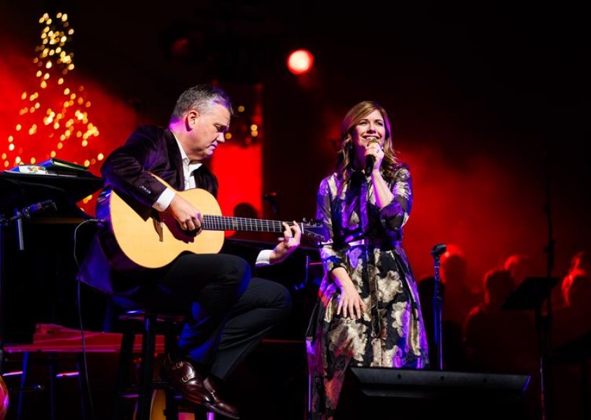 Keith and Kristyn Getty sitting down on chairs on a Stage performing. Keith holds an acoustic guitar and Kristyn is holding a Microphone.x